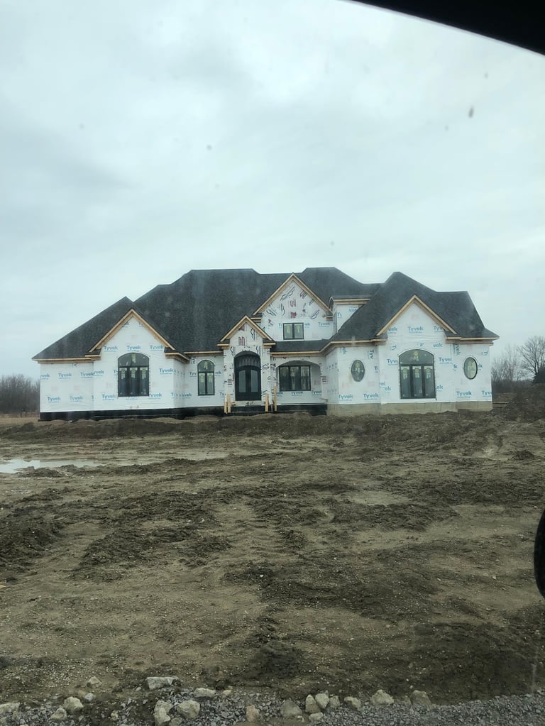 Large two-story white brick house under construction with black roof and dark shutters on bare dirt lot under cloudy sky