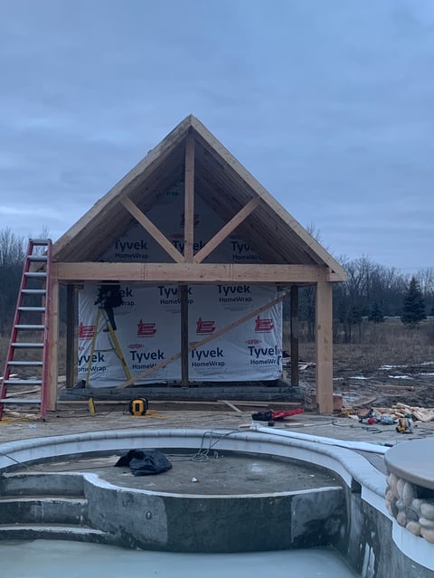 House frame under construction with Tyvek wrap, ladder, and construction materials, concrete foundation in foreground