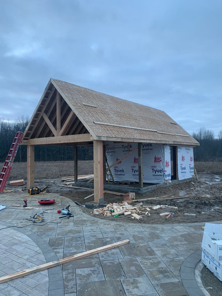 A timber-framed house under construction with pitched roof, Tyvek wrapped walls, and a paved foundation in progress