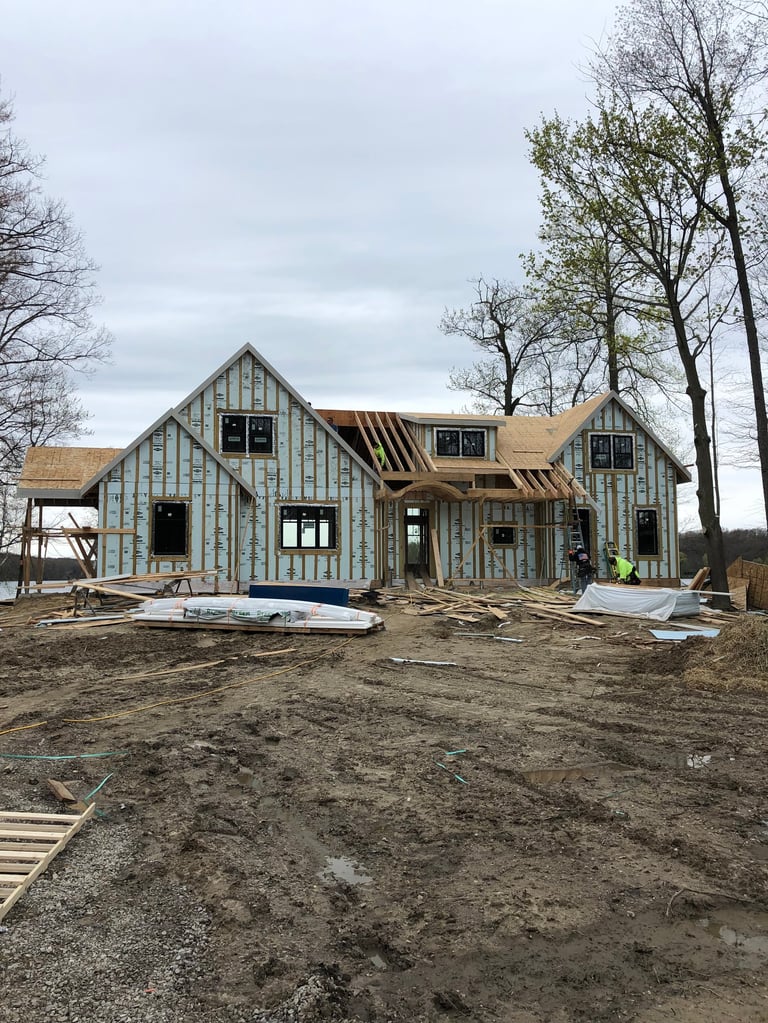 Modern farmhouse under construction with blue-gray exterior framing, exposed wood roofing, and barren construction site with surrounding bare trees