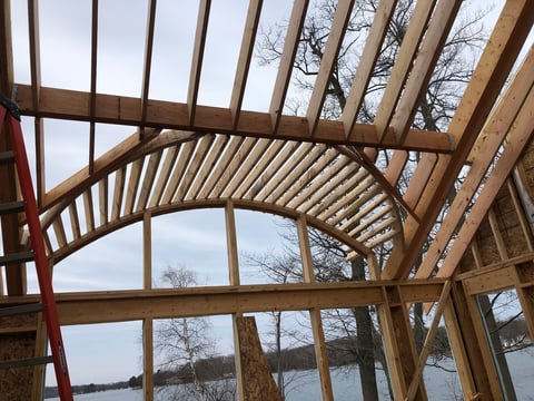 Wooden roof structure with curved beams and large windows overlooking a frozen lake and bare trees